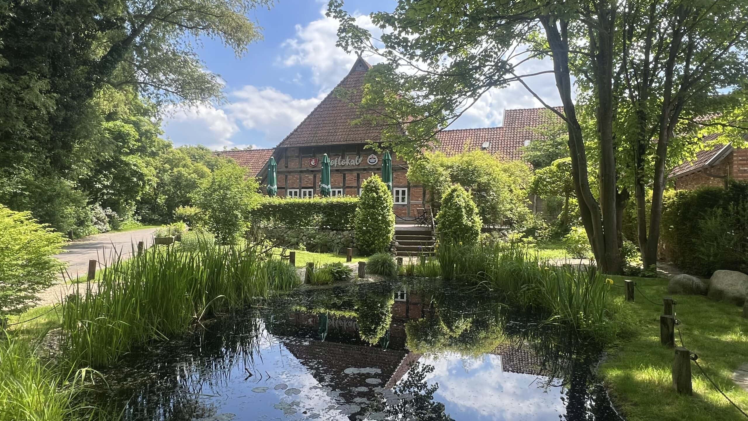 Fachwerkhaus mit Caféterrasse hinter Teich und Bäumen, umgeben von grüner, idyllischer Natur.